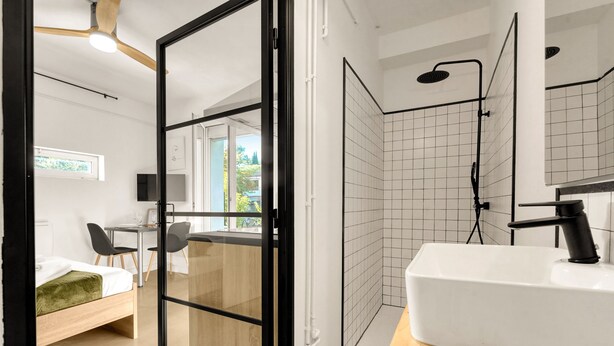 Ein modernes Badezimmer mit Dusche, Waschbecken und Toilette. Im angrenzenden Raum befindet sich ein Bett mit grüner Decke sowie ein Schreibtisch und ein Stuhl.