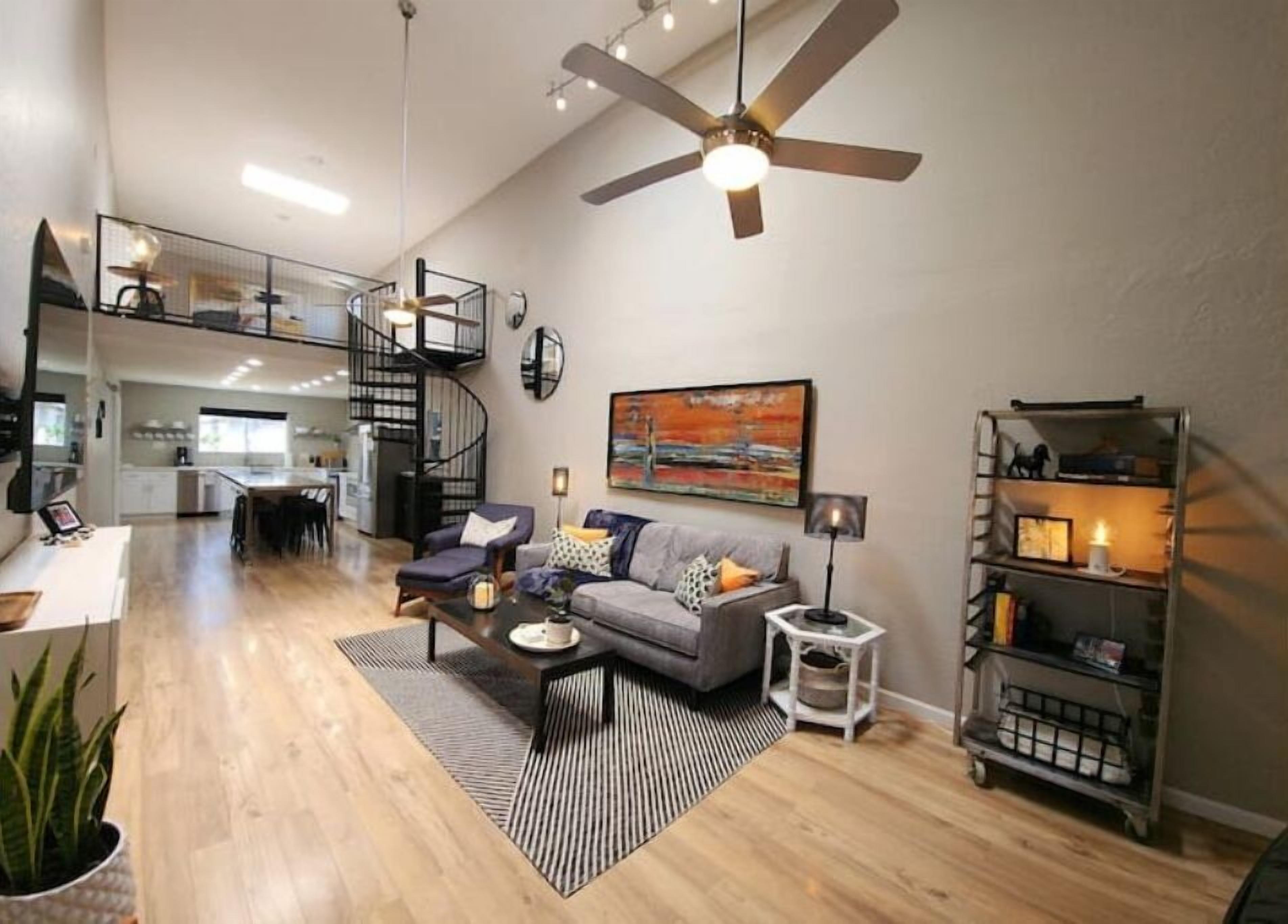 A spacious living area with a modern ceiling fan, a grey sofa with cushions, a coffee table, and a bookshelf.