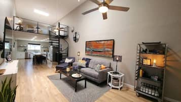 A spacious living area with a modern ceiling fan, a grey sofa with cushions, a coffee table, and a bookshelf.
