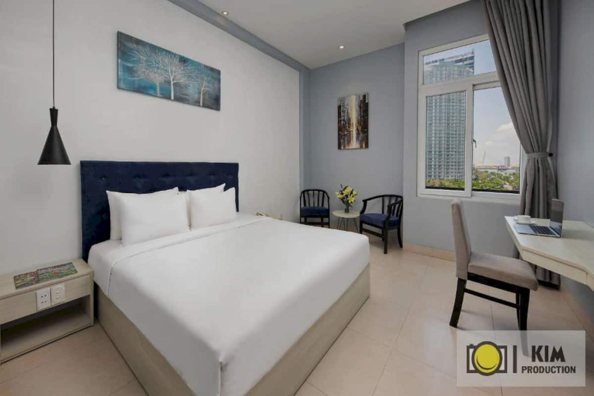 A modern hotel room with a large bed, a desk with a laptop, a chair, and a window with a city view.