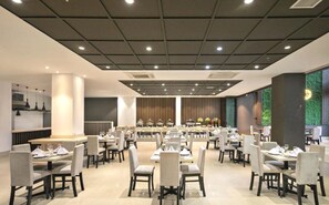 A spacious dining area with multiple tables and chairs, a central buffet, and a green wall with vertical lines. - Capital O 1200 Salem Riverside Hotel (Da Nang)