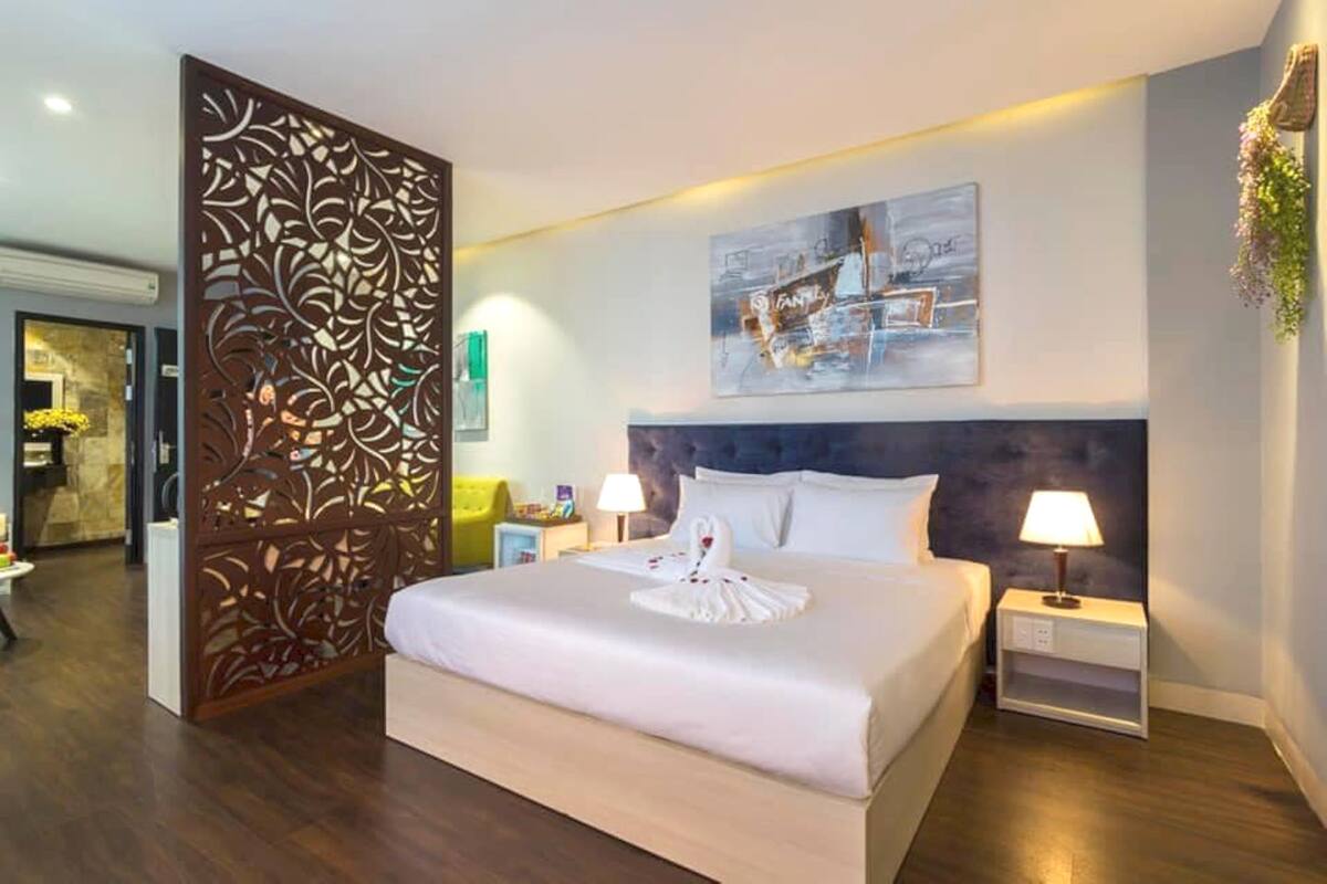 A hotel room with a large bed, a decorative partition, a painting on the wall, and a nightstand with a lamp.