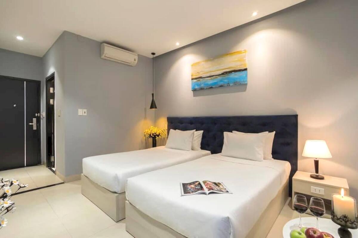A modern hotel room with two beds, a large painting on the wall, a bedside table with a lamp, and a small table with a candle and fruit.