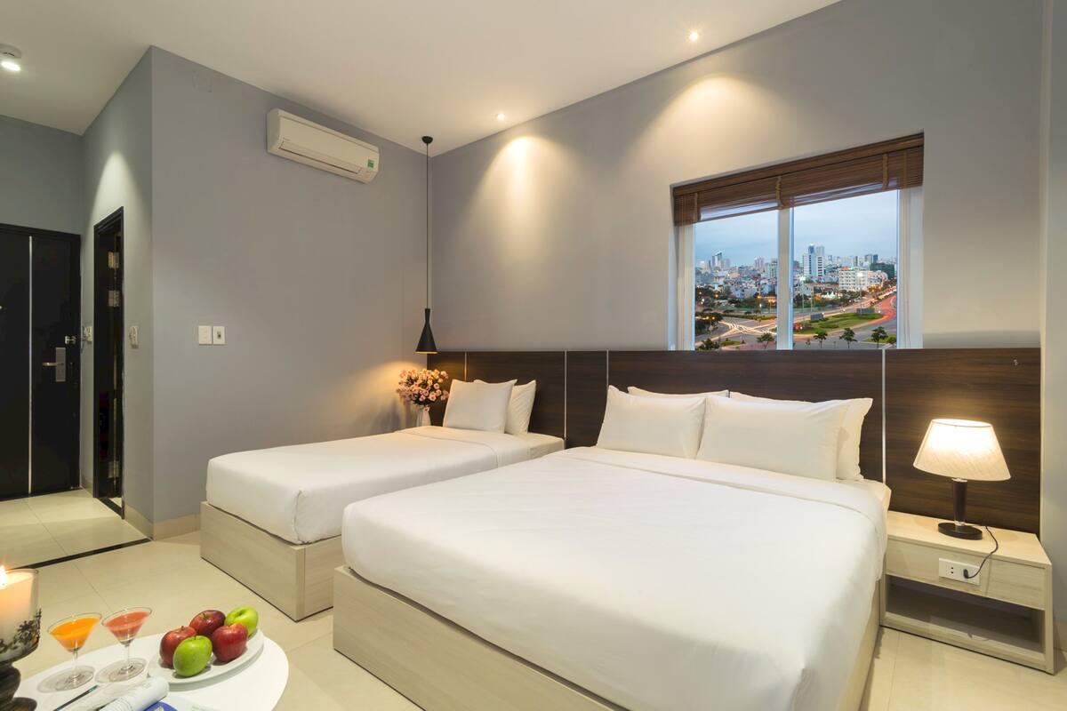 A hotel room with two beds, a city view, a bedside table with a lamp, and a small table with fruit and drinks.