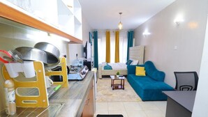 A compact living space with a kitchenette, a bed with blue and yellow bedding, a blue sofa, and a small table. - Songeza Furnished Accomoda-super Studio,embakasi (Nairobi)