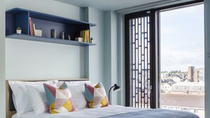 A modern bedroom with a bed, pillows, a blue shelf, and a view through a window. - Coleman Court Summer Accommodation (Cork)