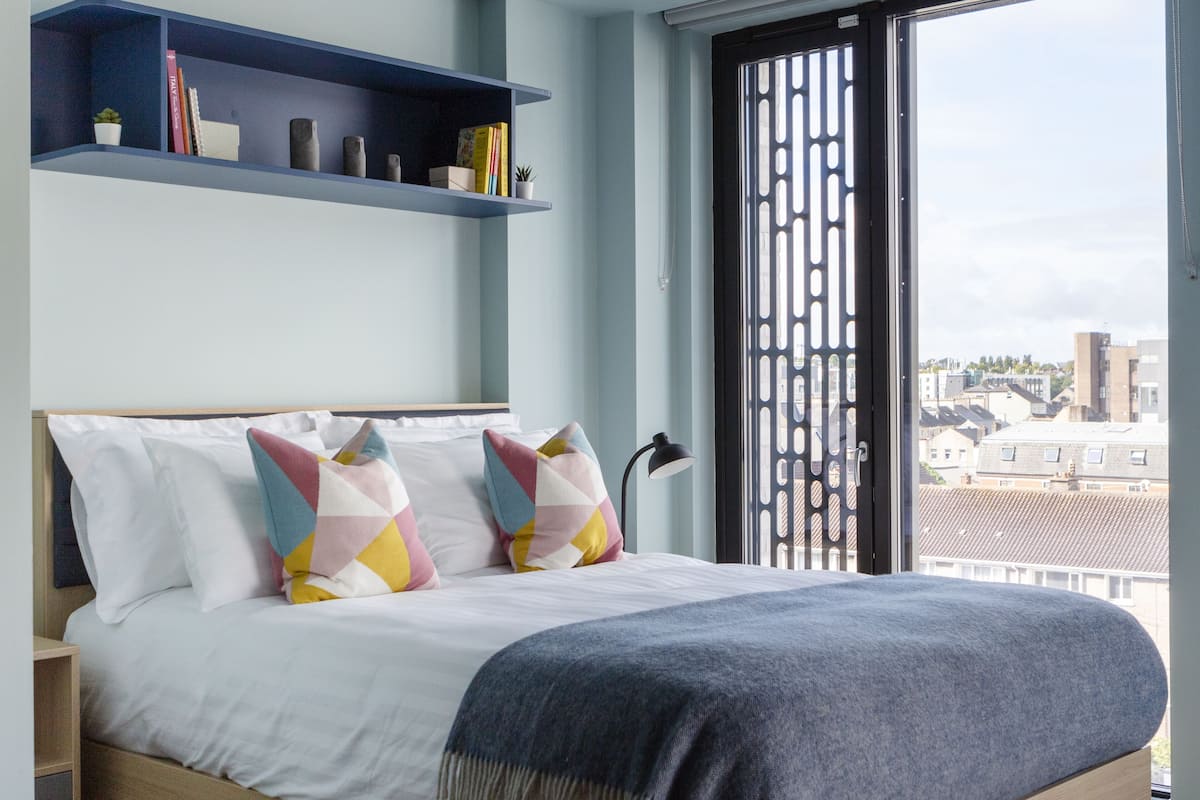 A modern bedroom with a bed, pillows, a blue shelf, and a view through a window.