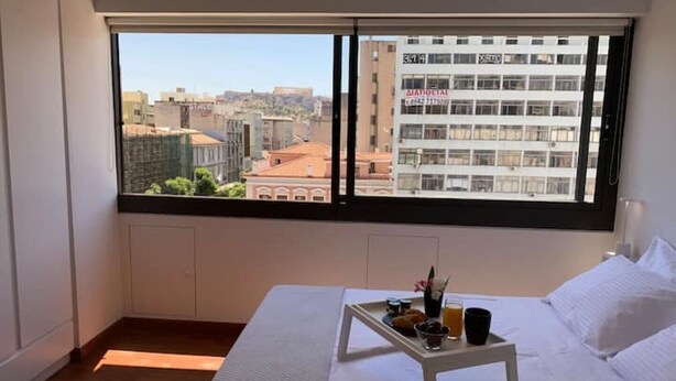 A hotel room with a large window offering a city view, a bed with white linens, a small bedside table with a tray of breakfast items, and a white nightstand. - Athens Market Iconic Apartment, Amazing view of the Acropolis (Athens)