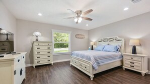 Een slaapkamer met een bed, nachtkastjes, een dressoir, een plafondventilator en een raam met uitzicht op groen.