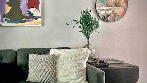 Ein Wohnzimmer mit einem dunkelgrünen Sofa, einem weißen Strick-Zierkissen, einer Zimmerpflanze, einem runden Gemälde und einem kleinen Tisch mit Buch und Vase.