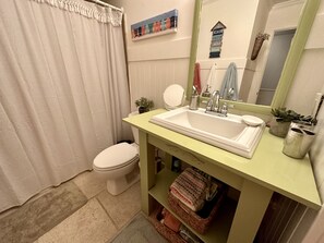 Un baño con inodoro blanco, lavamanos con encimera verde, espejo y estante con artículos de aseo.