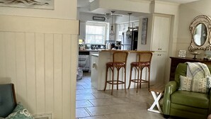 Una sala de estar con un sofá verde, un sillón azul y una pequeña mesa. Se observa una cocina al fondo con un refrigerador, un lavamanos y una ventana.