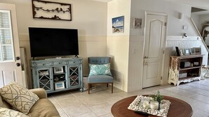 Una sala de estar con un sofá color beige, un sillón azul, una mesa de centro de madera con objetos decorativos, un televisor y un mueble con marcos de fotos.