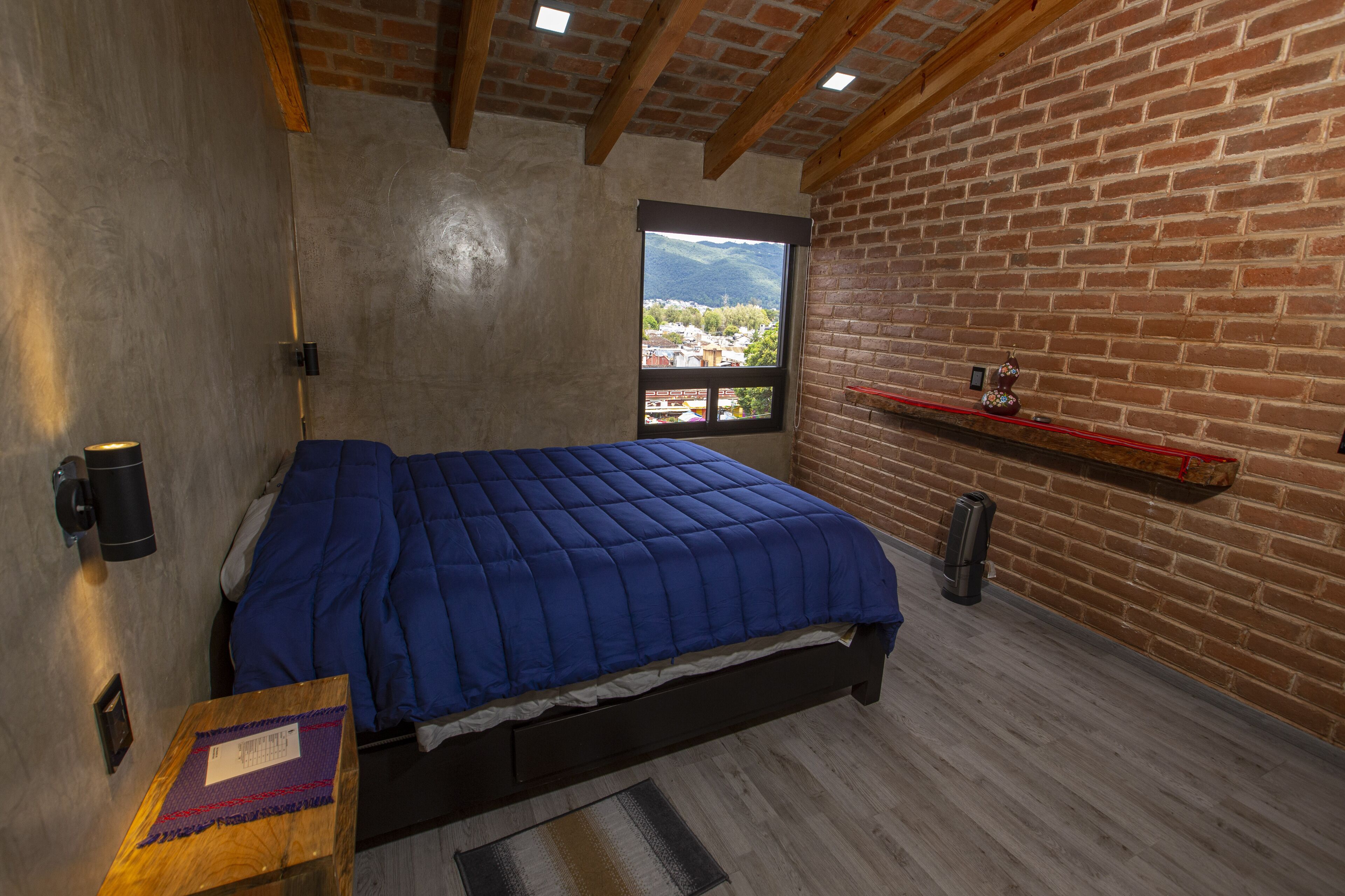 A bedroom with a bed, a window with a view, a brick wall, and a shelf with a vase.