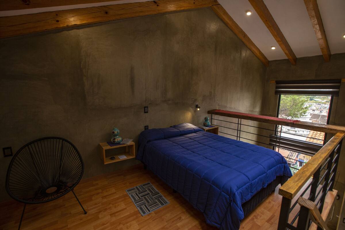 A bedroom with a wooden ceiling, a bed with a blue bedspread, a chair, a nightstand, and a window with a view.