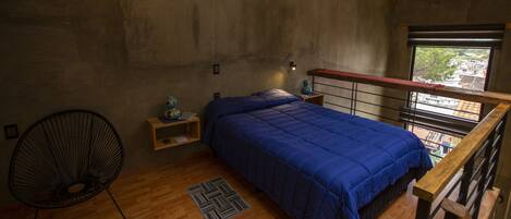 A bedroom with a wooden ceiling, a bed with a blue bedspread, a chair, a nightstand, and a window with a view.