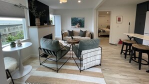 Un salon moderne avec un canapé, une table basse et des fauteuils, un coin repas avec une table et des tabourets, et une cuisine en arrière-plan.
