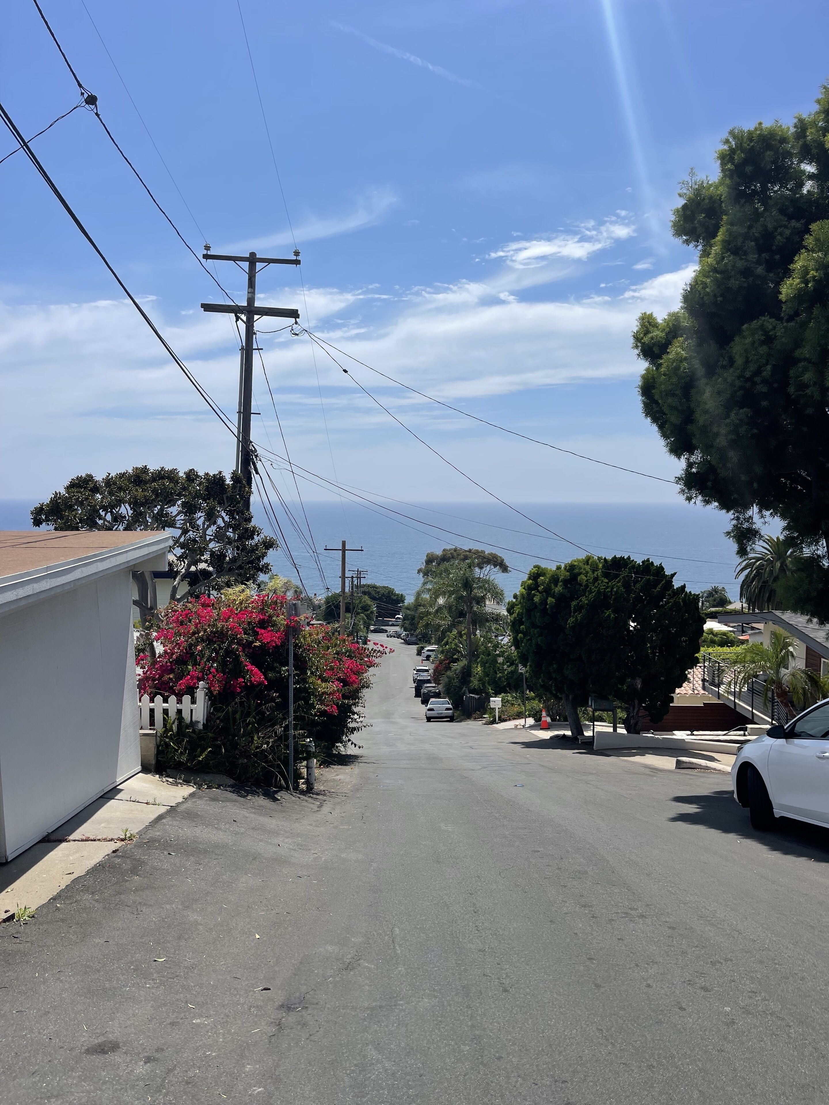 A residential street with houses, a white car parked on the right, and a clear view of the ocean in the distance.
