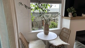 Une pièce avec une table ronde blanche, deux chaises en osier dotées d’assises blanches et moelleuses, une plante en pot posée sur la table, et une fenêtre donnant sur une clôture en bois et de la verdure à l’extérieur.