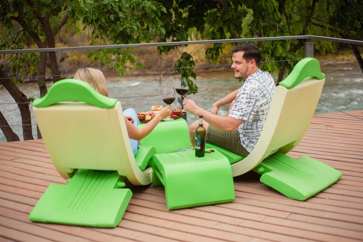Dos personas sentadas en sillones verdes sobre una terraza de madera junto a un río.