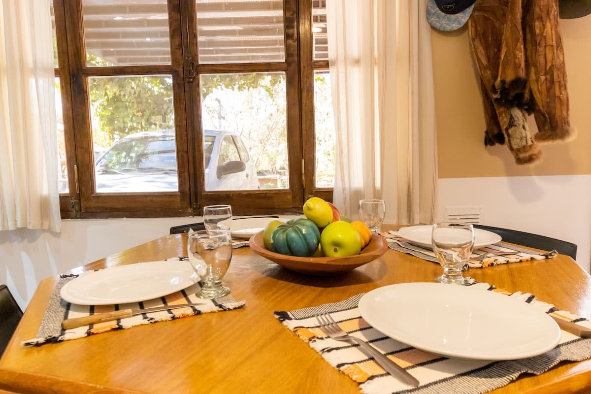 Una mesa de comedor servida con platos, cubiertos, un frutero y una ventana con persianas.