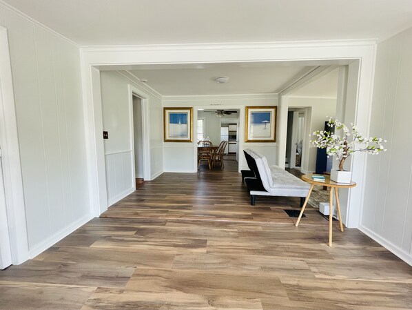 A hallway with a wooden floor, white walls, and a seating area with a sofa and a small table. There are framed pictures on the walls and a potted plant on the table.