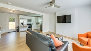 Una sala de estar moderna con un sofá de cuero negro, un sillón naranja, un televisor de pantalla plana montado en la pared y un ventilador de techo.