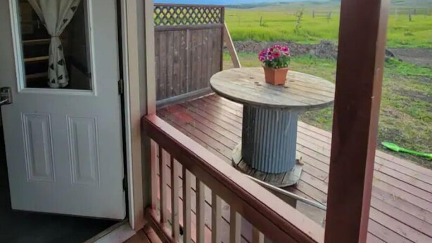 Una terraza de madera con una mesa redonda y plantas en macetas, con vistas a un campo verde.
