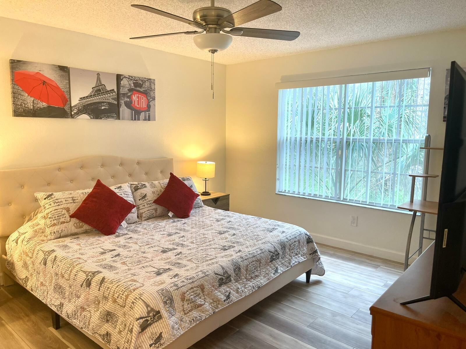 A bedroom with a bed, a ceiling fan, a window with a view of trees, and artwork on the wall.