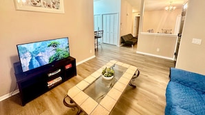 A living room with a flat-screen TV, a glass-top coffee table, a blue sofa, and a framed picture on the wall. - Grand Family 3BD Condo Apartment Near Disney Parks 5 (Kissimmee)