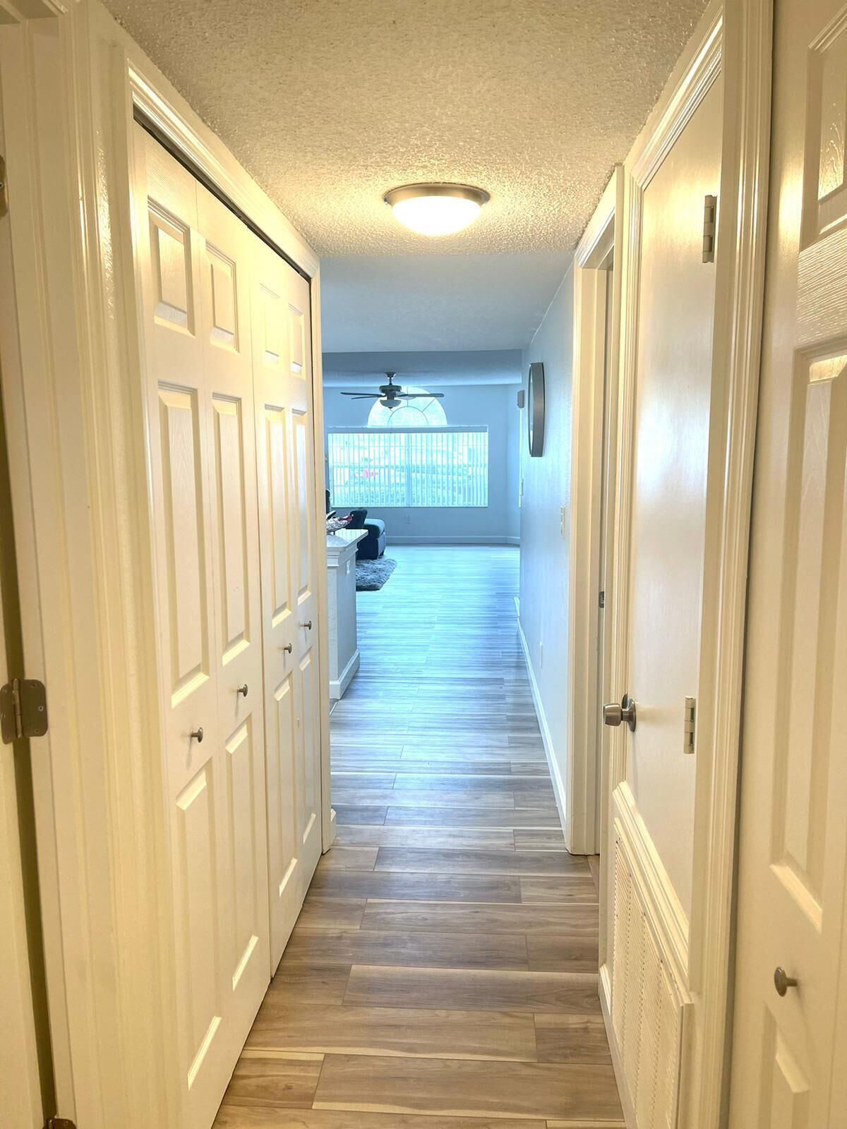 A hallway with white doors, a ceiling fan, and a view to a room with a window and a ceiling fan.