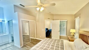 A bedroom with a bed, a ceiling fan, a nightstand with a lamp, a window with blinds, and a door leading to another room.