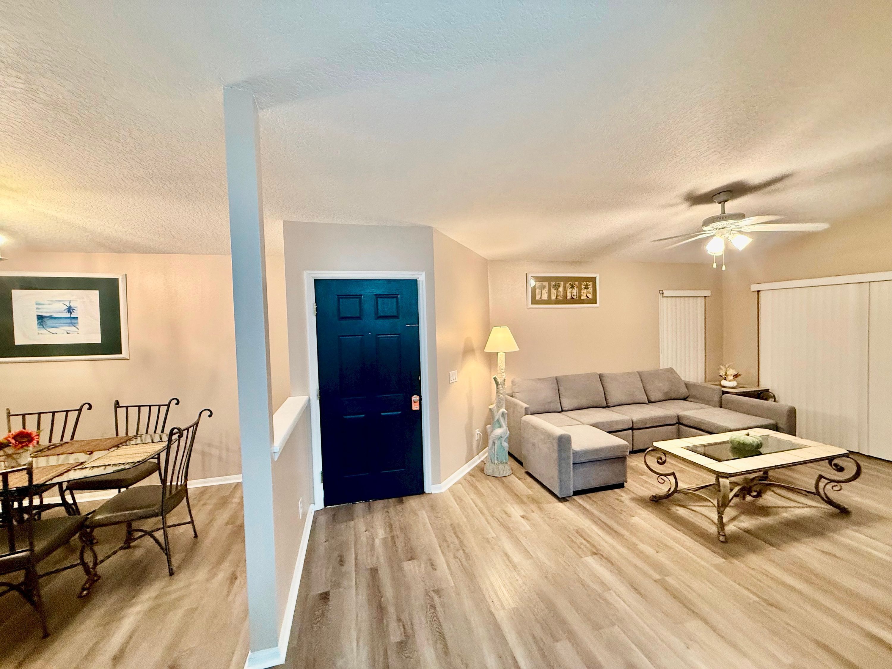 A living room with a sofa, a dining table with chairs, a ceiling fan, and a door.