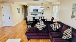 A living room with a purple sofa, wooden flooring, a dining area with a kitchen in the background, and a white door. - Grand Premier 3BD Condo Apartment Near Disney Parks 6 (Kissimmee)