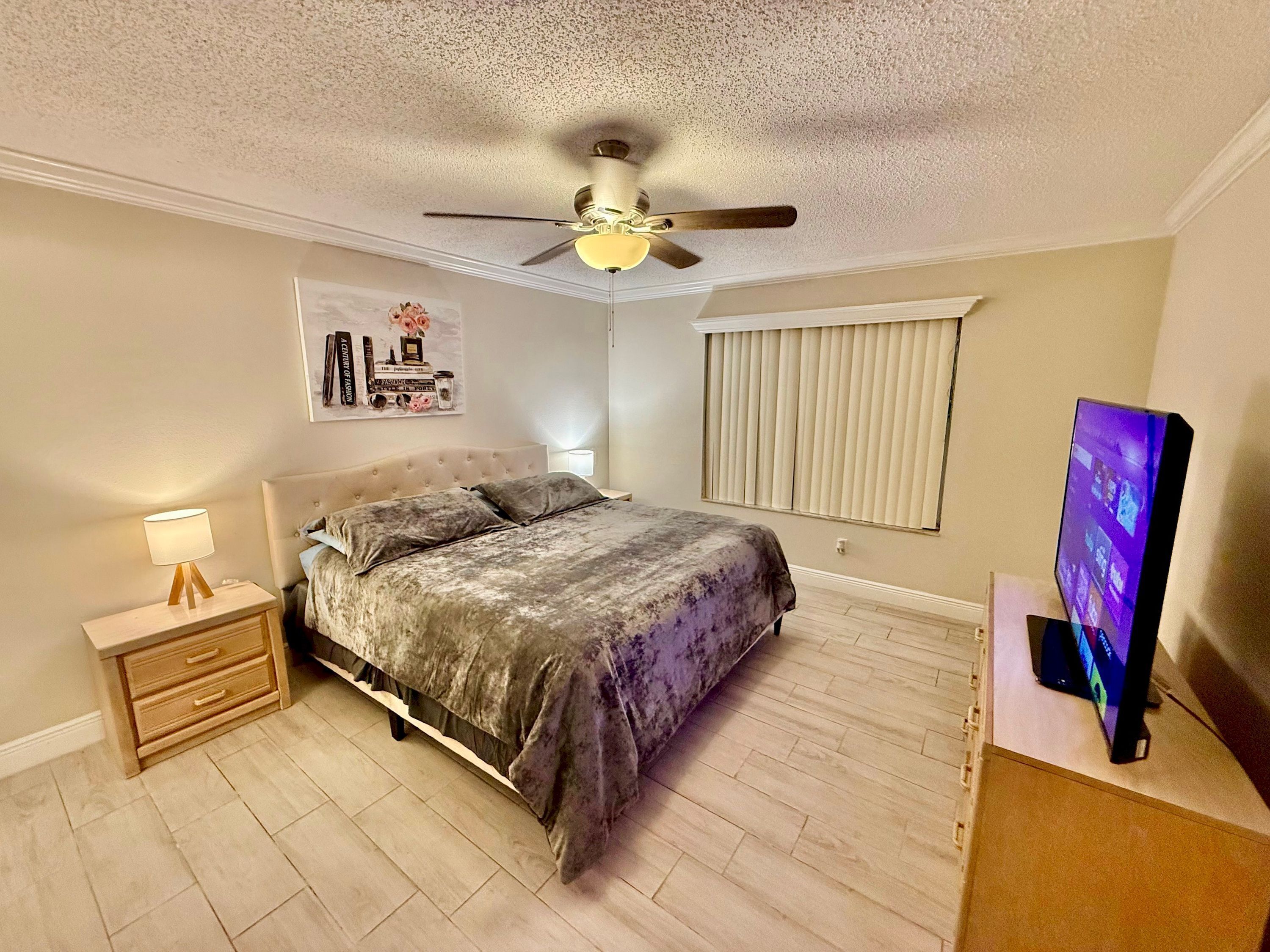 Une chambre à coucher comprenant un lit, une table de chevet, une lampe, un ventilateur de plafond, des stores et un téléviseur.