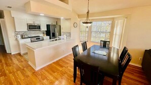 A modern kitchen with a central island, dining table with chairs, and a large window with blinds. - Grand Premier 3BD Condo Apartment Near Disney Parks 6 (Kissimmee)