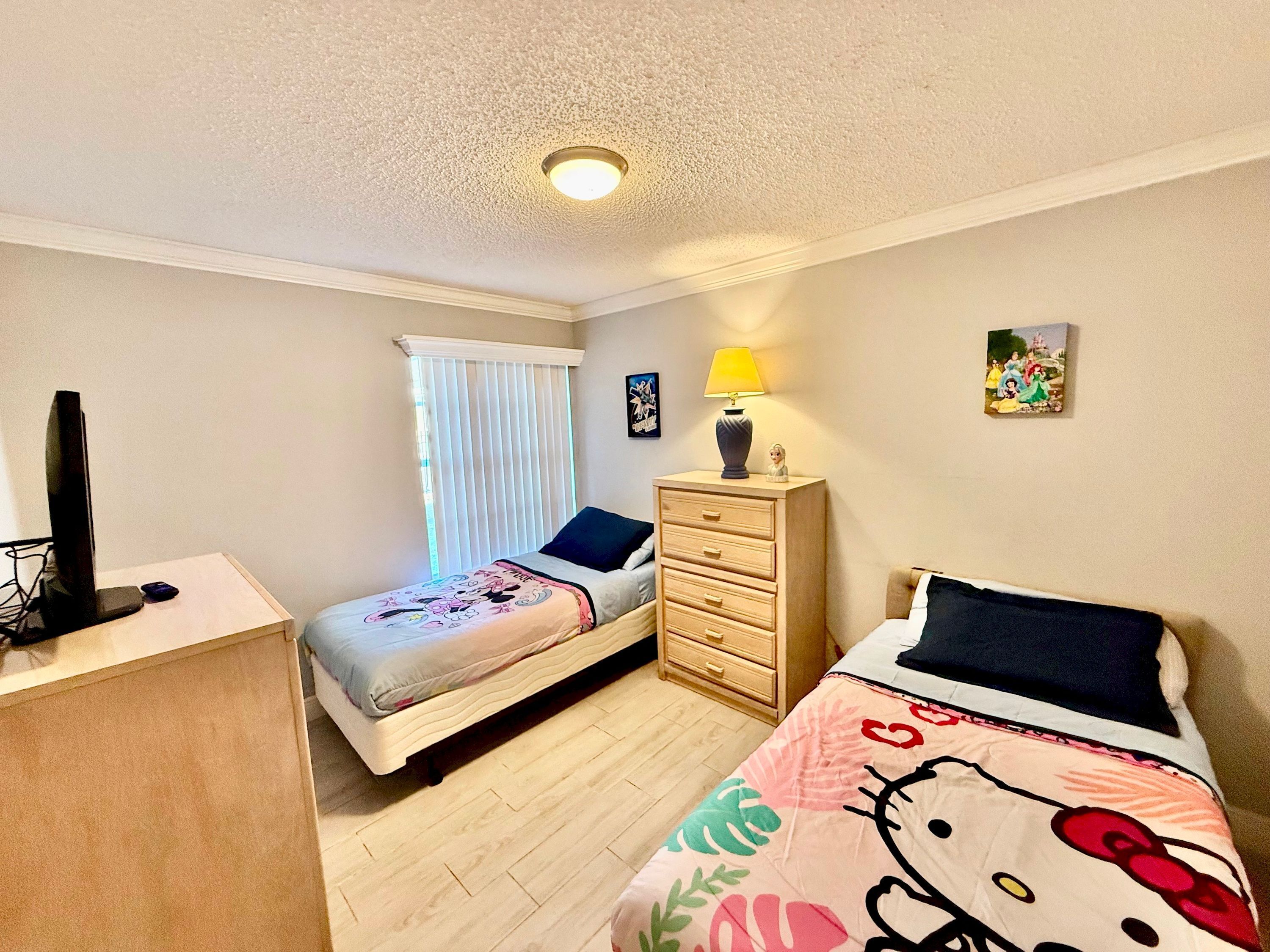 A bedroom with two beds, a desk, a dresser, a lamp, and a window with blinds.