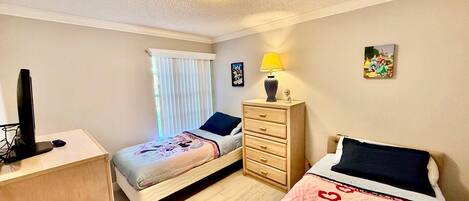 A bedroom with two beds, a desk, a dresser, a lamp, and a window with blinds.