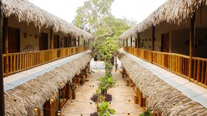 Ein Korridor mit reetgedeckten Zimmern, einem zentralen Gang mit Pflanzen und Blick auf einen Baum.