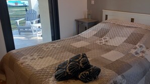 Ein Bett mit einer gesteppten Decke und einem gemusterten Kissen, ein Nachttisch mit Lampe und Blick auf eine Außenpatio-Terrasse.