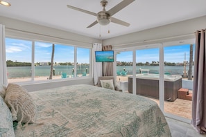 Ein Schlafzimmer mit einem großen Bett, einem Deckenventilator und einem Fernseher. Es gibt ein großes Fenster mit Blick auf das Wasser und eine Whirlwanne draußen.