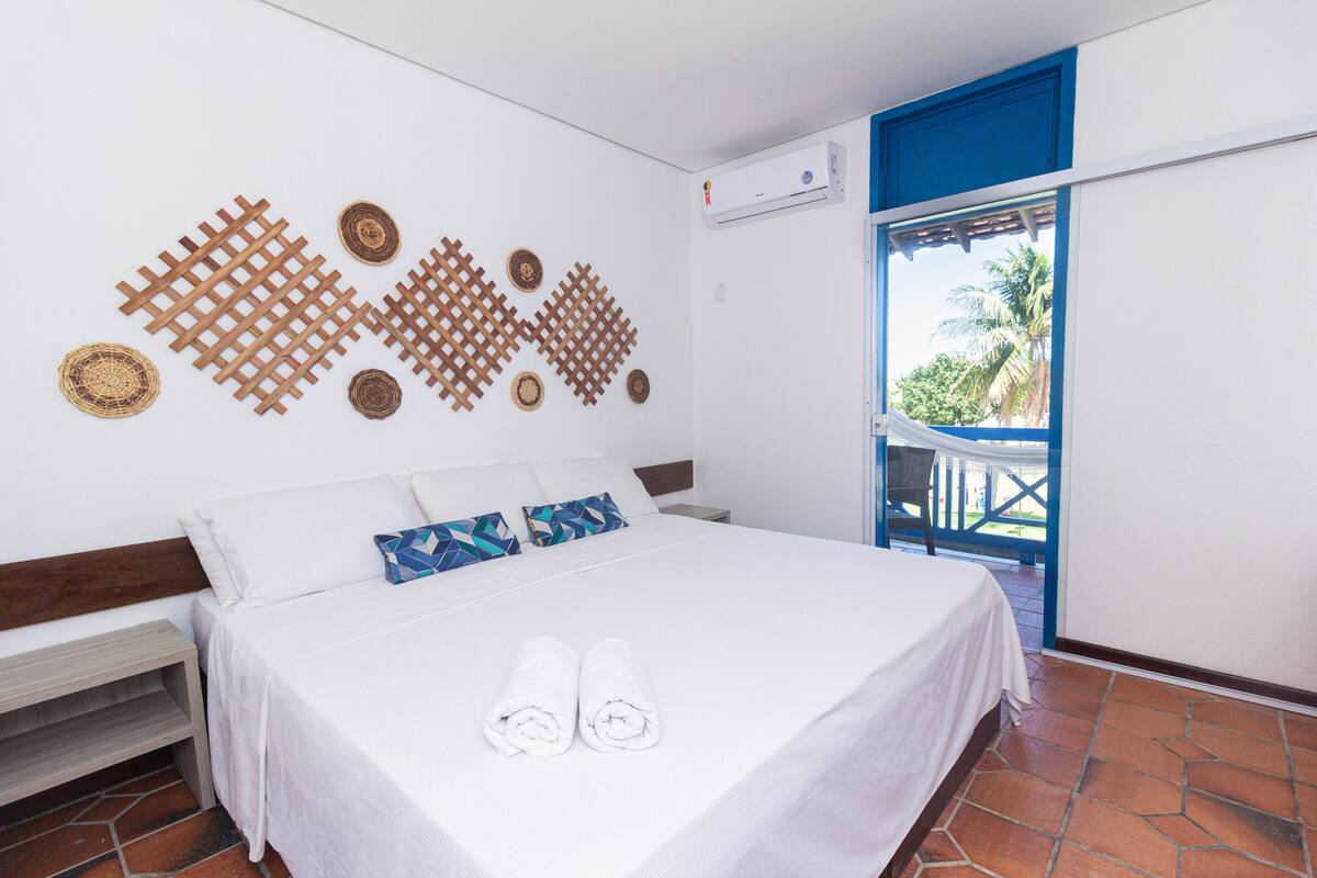 A neatly made bed with white linens and rolled towels, a wooden headboard, and a wall decorated with geometric patterns.
