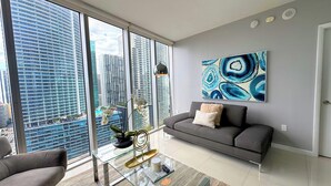 A modern living room with a grey sofa, a glass coffee table, and a view of tall buildings through large windows. - Icon Brickell Condo - We Host (Miami)