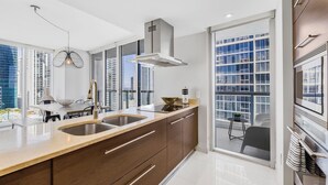 A modern kitchen with a large island, stainless steel appliances, and a view of the city through large windows. - Icon Brickell Condo - We Host (Miami)