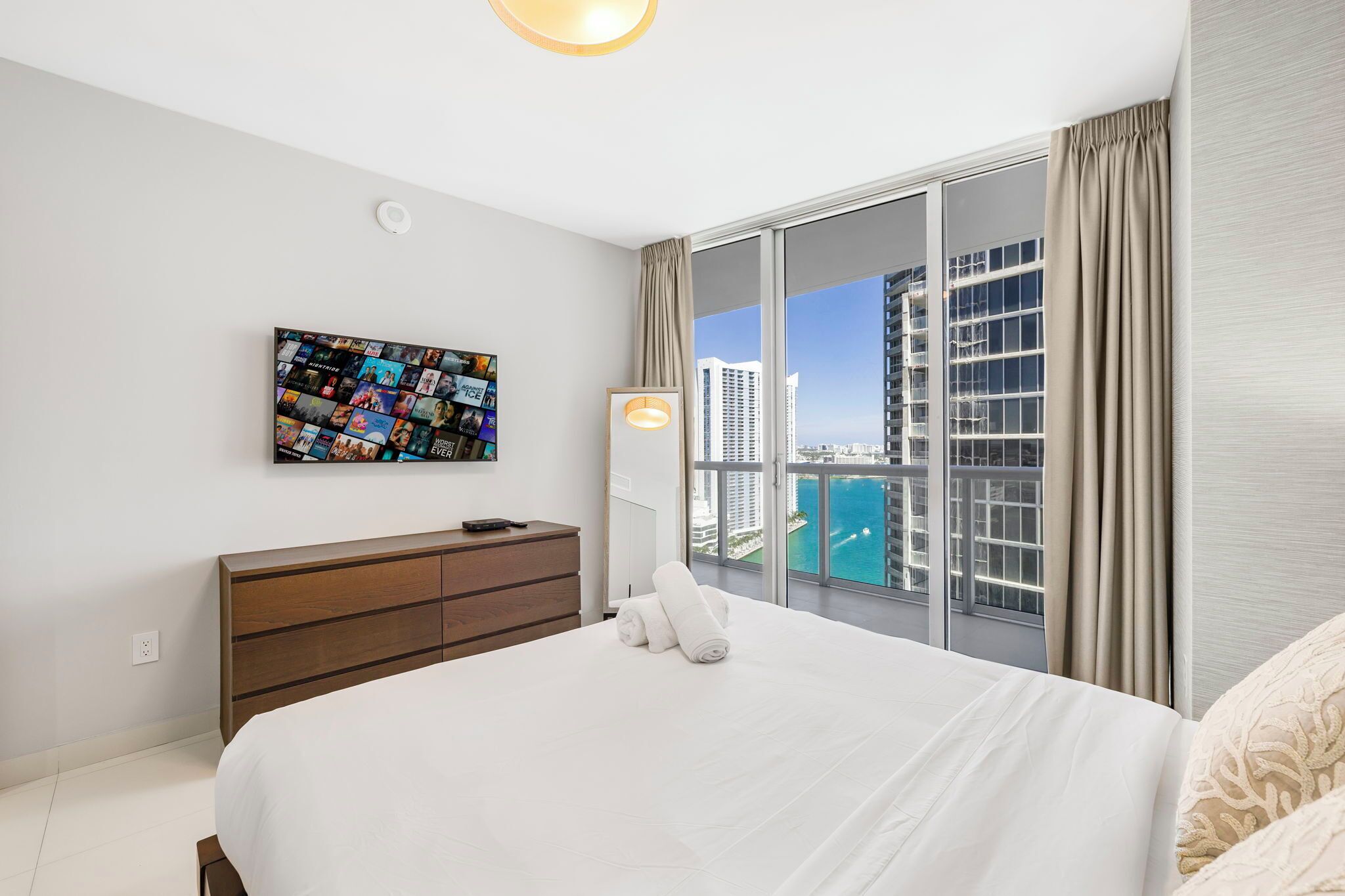 Une chambre d’hôtel dotée d’un grand lit, d’une commode en bois, d’un téléviseur fixé au mur qui affiche un montage de photos et d’une grande fenêtre donnant sur un paysage urbain et l’eau.