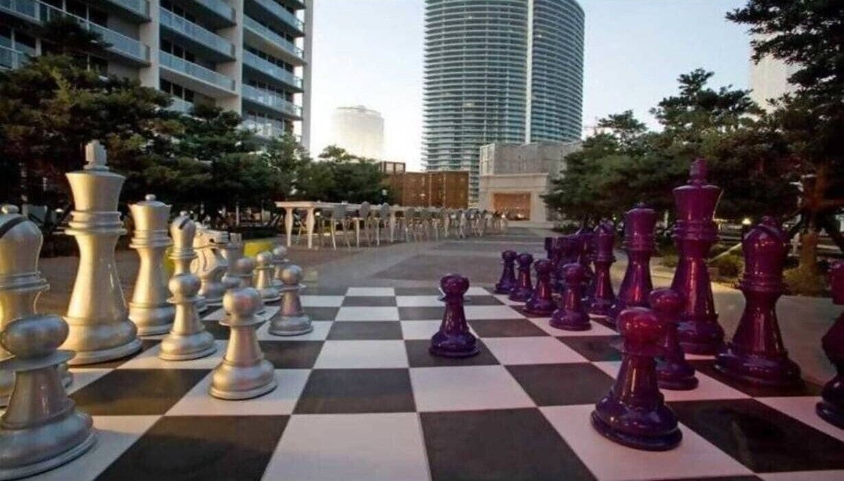 A large outdoor chessboard with chess pieces set up in a public space, surrounded by trees and buildings.