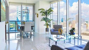 A modern living room with a glass coffee table, a sofa, and a dining area with chairs. There is a large window with a city view. - Icon Brickell Condo - We Host (Miami)