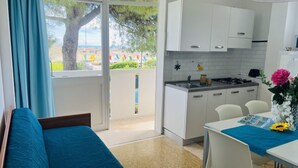A compact kitchen with white cabinets, a dining table with chairs, and a view of a tree and a beach through the window. - Cozy Apartment Facing the sea (San Michele al Tagliamento)