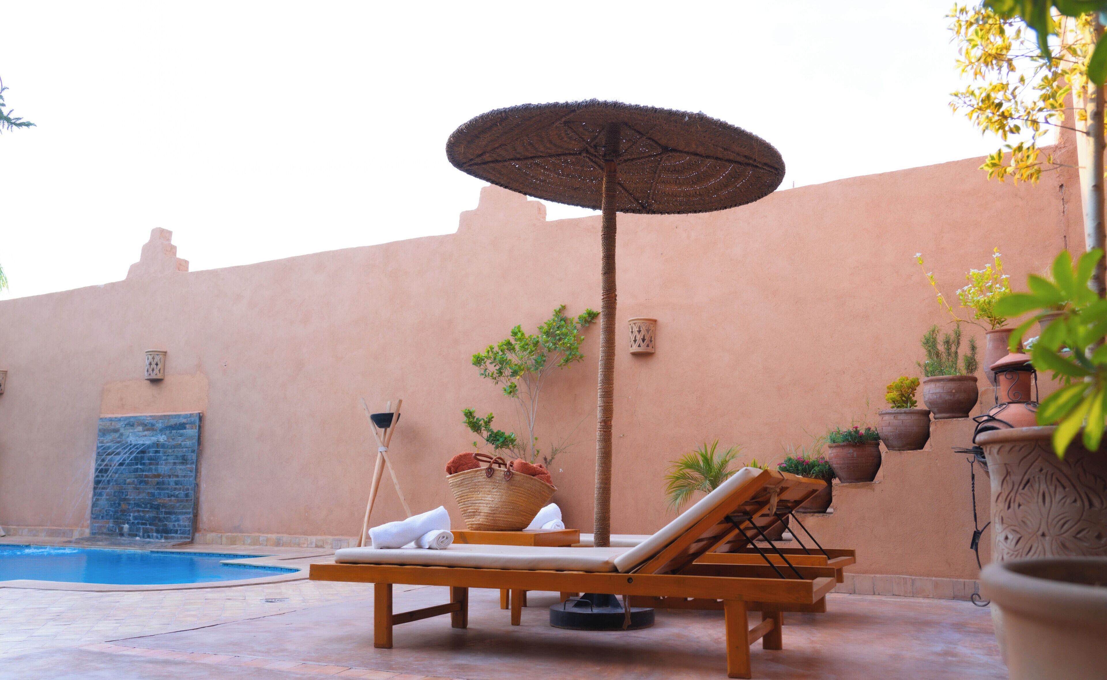 Un espace aménagé au bord de la piscine, avec des chaises longues, un parasol, une petite table sur laquelle se trouve un panier, et des plantes en pot.