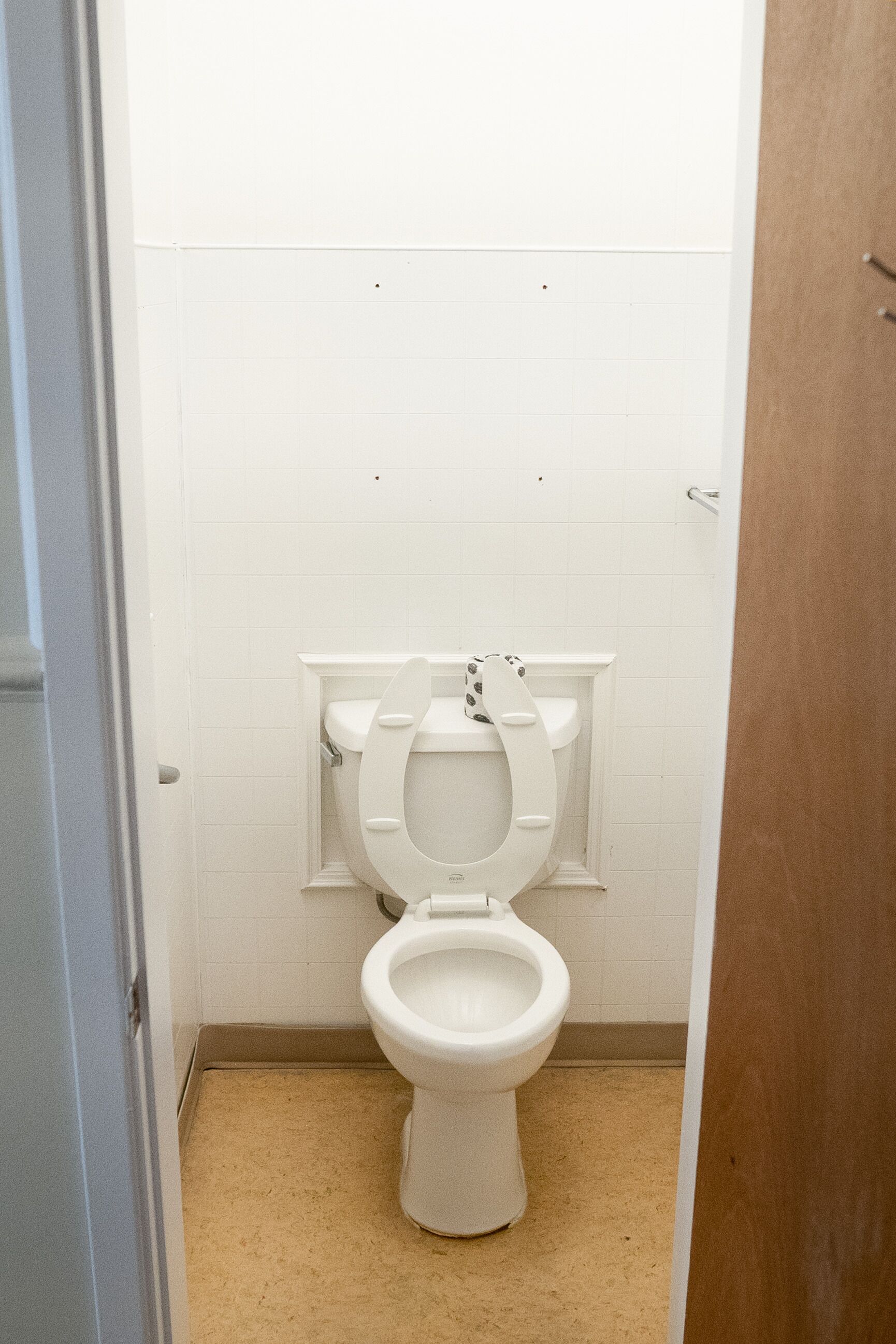 A small, white toilet in a narrow bathroom with white tiled walls and a beige floor.